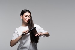 positive young woman using hair straightener isolated on grey