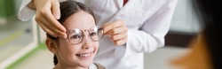 oculist trying glasses on smiling girl near blurred mother in optics store, banner