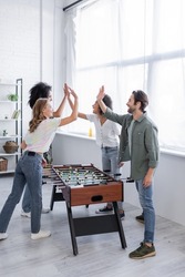 happy interracial friends giving high five near table football