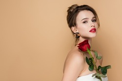 pretty young woman in strapless top holding red rose and looking at camera isolated on beige