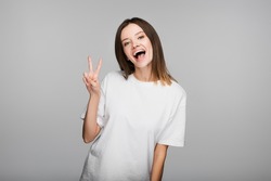 thrilled woman in white t-shirt showing victory sign while looking at camera isolated on grey