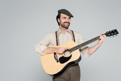 smiling man with mustache in retro style clothing and hat playing acoustic guitar isolated on grey