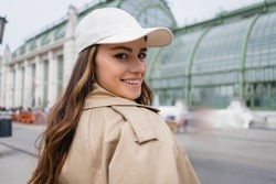 happy young woman in beige trench coat and baseball cap
