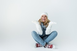 cheerful blonde woman in knitted hat and winter outfit sitting and gesturing on grey