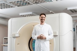 happy radiologist in white coat standing near computed tomography scanner and looking at camera