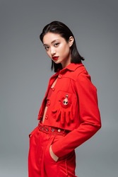 asian woman in red jacket decorated with brooch and glove standing with hands in pockets isolated on grey
