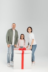 Full length of smiling parents and preteen daughter looking at camera near big gift box during child protection day celebration on grey background