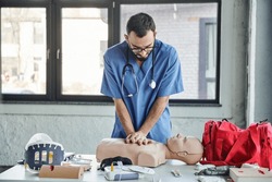 young paramedic in blue uniform and eyeglasses practicing chest compressions on CPR manikin near defibrillator and first aid kit during medical seminar, life-saving skills development concept