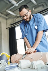 bearded professional paramedic in eyeglasses and blue uniform practicing chest compressions on CPR manikin near defibrillator and compressive bandages, critical skills development concept
