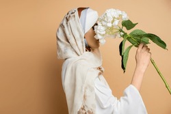 side view of modest muslim woman in headscarf and blouse obscuring face with white flower on beige