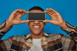 happy african american guy obscuring face with smartphone on blue backdrop, obscuring face