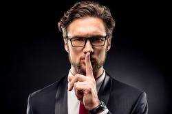 Handsome businessman in eyeglasses and suit gesturing for silence with finger isolated on black