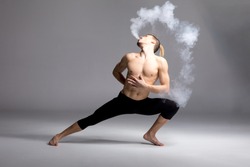Young shirtless man dancer vaping and blowing smoke on grey