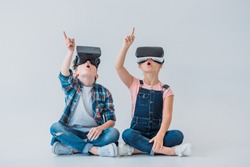 amazed kids using virtual reality headsets and pointing up with finger while sitting on the floor