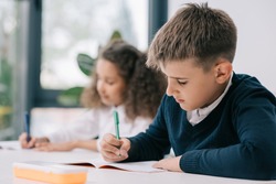 Concentrated schoolboy sitting at desk and writing in exercise book with classmate sitting behind