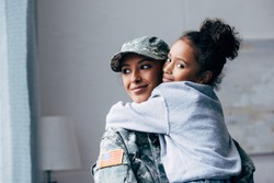 smiling african american soldier in military uniform holding daughter on arms at home