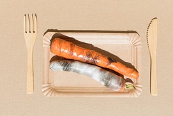 Creative food. Carrots wrapped in polyethylene plastic on the beige paper background. Immature, tasteless or inedible food concept. Copy space. Flat lay.