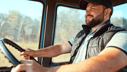 View on cabin on young male with beard sits driving his tractor and drives through field and smiles toothily and laughs. Happy farmer driver. Cultivation of farmland. Plowing agricultural technology.