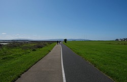 Walking & bicycle path from Galway to Salthill, Ireland with views of the grassy sports field and hills across the bay, with silhouetted figures walking