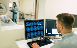 Back view photo of a male doctor sitting near the computer and looking on patient in magnetic resonance imaging machine