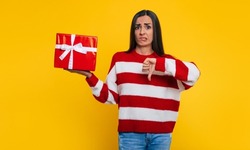 Close up photo of an upset young brunette woman while she shows the present box in hand and makes thumb down with unhappy face isolated on yellow wall