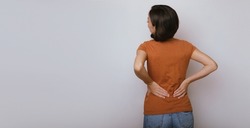 Spinal cord problems on woman's back. Chronic back pain. Young brunette woman is holding her lower back, while standing and suffering from unbearable and severe pain. Spine osteoporosis.