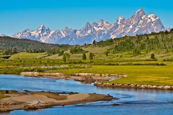 Grand Teton mountain range, partly covered in snow, with green forest, field, and small river in the foreground. It was taken on a sunny day in summer at the gateway to Grand Teton national park.