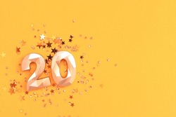 Golden number twenty and stars confetti scattered on a yellow background. Festive glittering compisition.