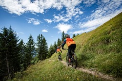 Two mountain bikers ride on the trail against the backdrop of incredible mountains at dawn. Two cyclists on a background of blue sky in an orange jersey
