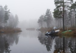 Fog. A few boats lie on the shore of the lake near some birch trees.