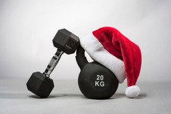 A large black Kettlebell wearing a red  Santa hat with a dumbbell leaning against it on a plain light gray background 