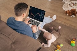 Businessman, crypto trader investor and young father working from home, using laptop with stock market app for cryptocurrency financial stock market analysis while his toddler baby playing nearby