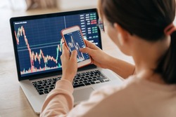 Woman crypto trader investor broker using smartphone app and laptop executing financial stock trade market trading order to buy or sell cryptocurrency. Selective focus on hands and cellphone screen