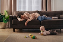 Young tired mother suffering from lack of sleep, sleeping on sofa while her little infant baby is playing on floor. Exhausted mom experiencing postnatal depression, does not want to play with her son