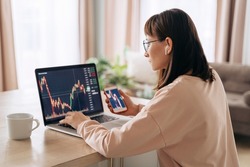 Young business woman trader analyst looking at laptop monitor, holding smartphone. Investor broker analyzing indexes, trading online investment data on cryptocurrency stock market graph at home