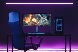 General view of home workplace of pro gamer with professional gaming setup on desktop. Modern powerful PC full RGB light inside, display with shooter game, armchair. Gaming studio of cyber sportsman