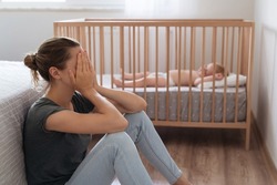 Side view of mother covering face crying from exhaustion sitting on floor next to baby crib while child napping, can't handle difficulties of maternity, having postnatal depression symptoms