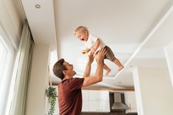 Young dad throwing excited laughing baby boy up in air and catching, amusing kid and having fun together after returning home from work while mother cooking dinner. Active play at home
