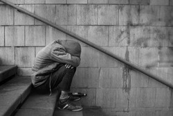 Drunk homeless unhappy man covered his face with his hands and sits on the stairs in the underpass, black and white photo.