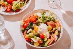 Healthy summer salad with fresh tomatoes, burrata cheese, olives and leafy greens on table with shadows, sunny mood, top view