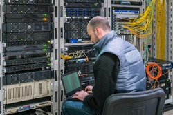 A man with a laptop sits in a server room. A technician works near the racks of a modern data center. The system administrator configures the computer hardware.