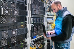 A man with a laptop stands in the server room. A technician works near the racks of a modern data center. The system administrator configures the computer hardware.