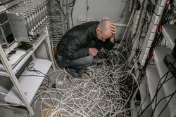 The man is in a plundered server room. A sad technician sits near a pile of wires and holds his head with his hands in an empty datacenter.