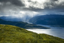 Dramatic storm clouds over the mountains and the Bergsfjorden fjord on Senja Island, with rain showers and ethereal rays of light piercing the tempest, as seen from the trail to Husfjellet Mountain.