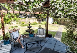 Young man lying down on patio lounge chair in outdoor spring flower garden in backyard porch of home happy smiling in zen with fountain, pergola canopy gazebo, table, plants, sofa