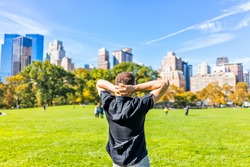 Back closeup of one happy young man with hands behind head, standing in Central Park in New York City during sunny autumn day with skyscrapers buildings and people