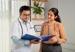 Asian Indian man or male physician or doctor wearing stethoscope and apron holding report file in hand and giving consultation to a smiling female or happy Woman patient. Medical, medicine, healthcare