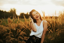 Gorgeous woman in a wheat field on a sunset background. A fashionable girl with long hair rejoices, laughs, enjoys life and summer, nature, happiness. Model in a hat in the forest.