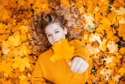 The girl lies in the autumn leaves and holds out one to the camera, a good autumn photo of a teenager
