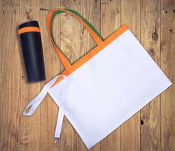 Blank white tote bag canvas for shopping with green and orange handle, white Lanyard Neck Strap for mockup. And Black thermal mug plastic travel with orange elements on wooden desk office. Over view.